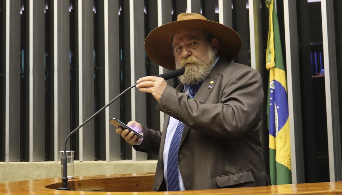 Nelson Barbudo, de chapéu, fala ao microfone na tribuna da Câmara dos Deputados, com a bandeira do Brasil ao fundo